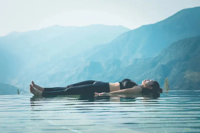 Woman laying in savasana floating on an ocean with mountain backdrop