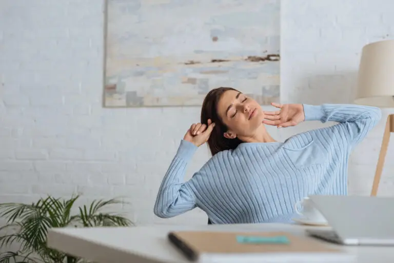 Woman sitting at desk, stretching with eyes closed, wearing light blue sweater