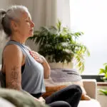 Woman sitting cross-legged on couch, eyes closed, hand over heart