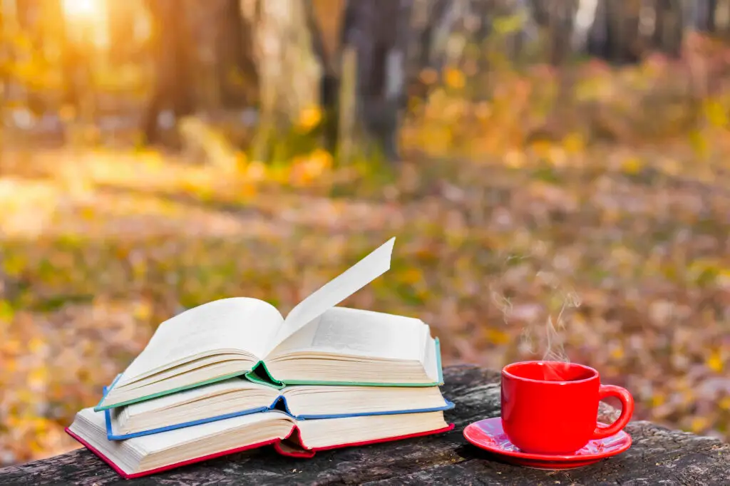 Open books and a red mug of coffee on a wooden table surrounded by autumn leaves in the woods.