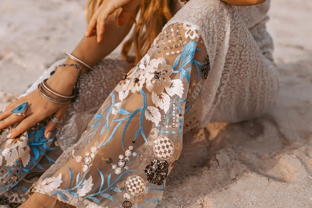 Bare feet and embroidered clothing with floral details resting on sand.