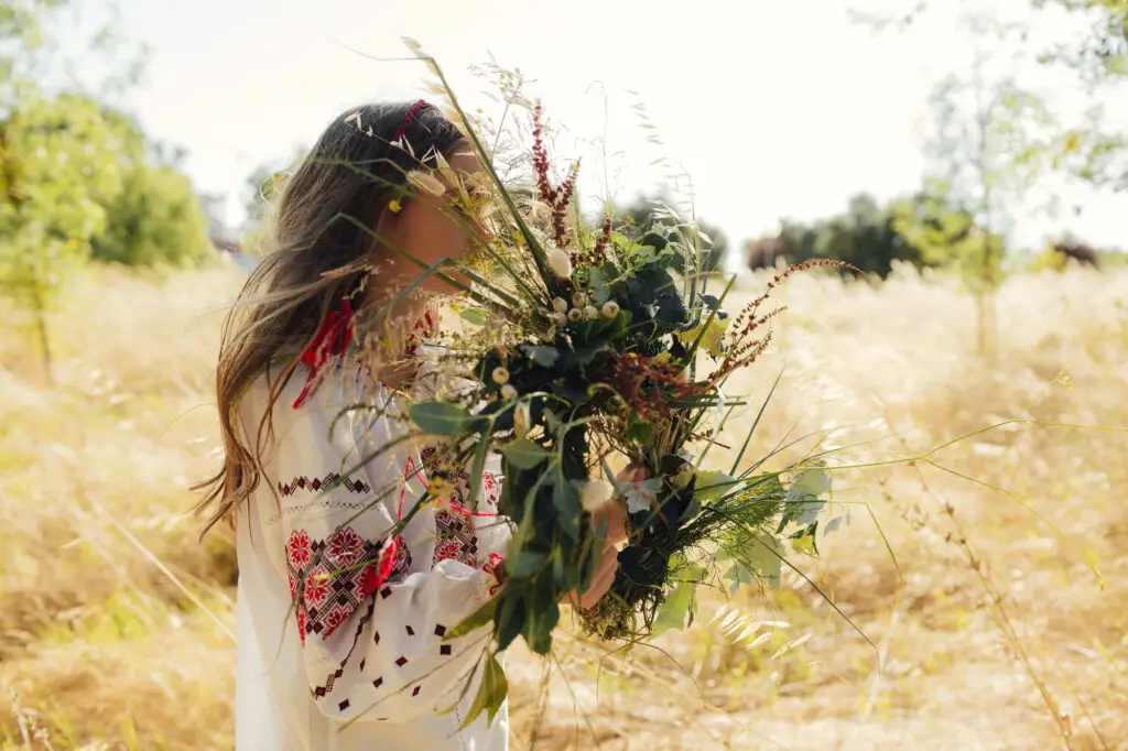 Woman carrying a large bouquet of wildflowers through a sunlit field.