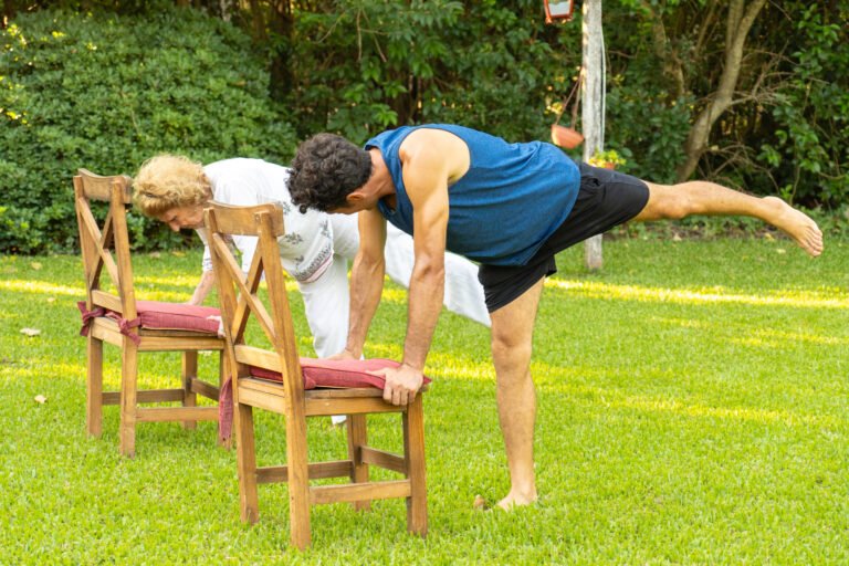 Older adults practicing a supported yoga balance pose with chairs outdoors.