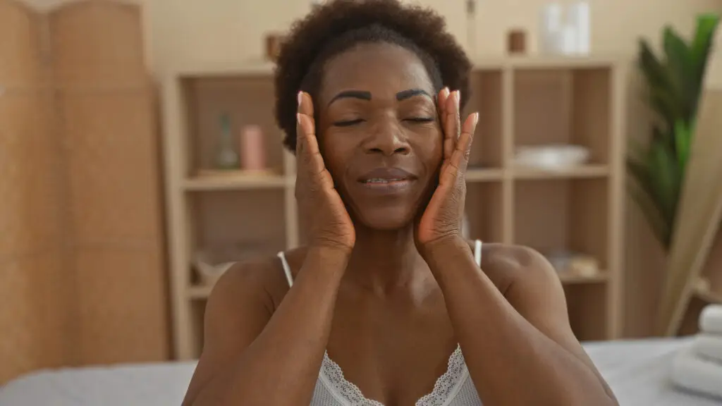 Woman with eyes closed and a gentle smile, resting both hands against her face in a moment of self-connection and calm.