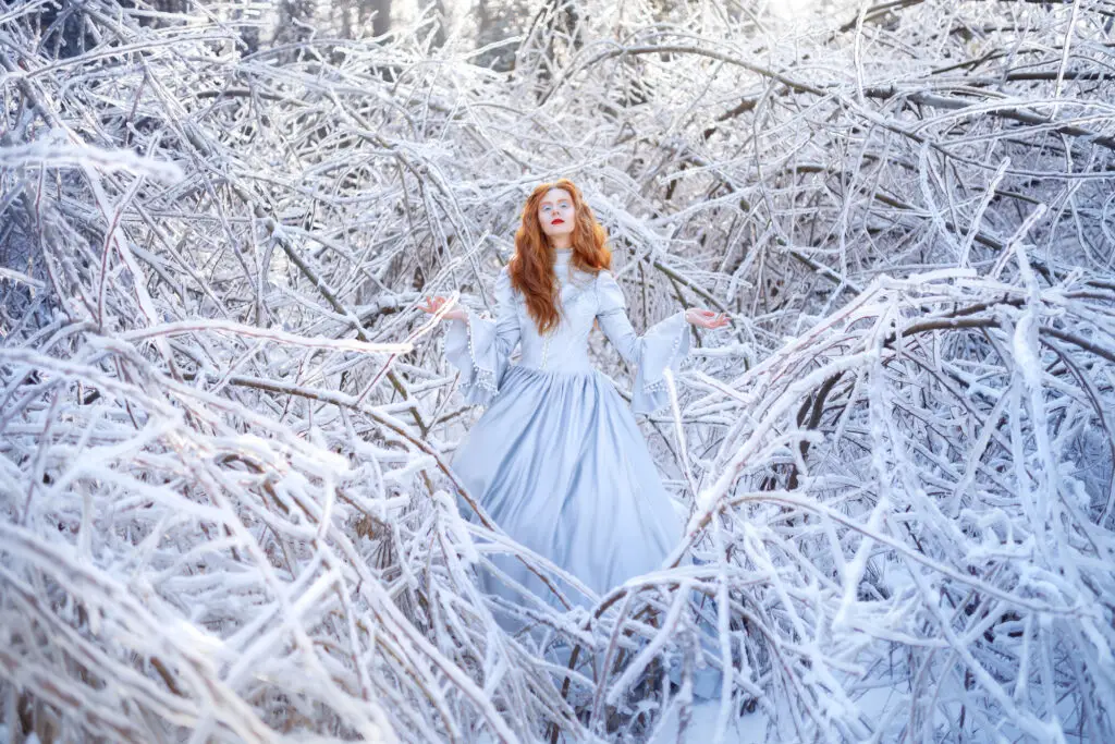 Red-haired woman in a pale blue gown surrounded by snow-covered branches in a winter forest.