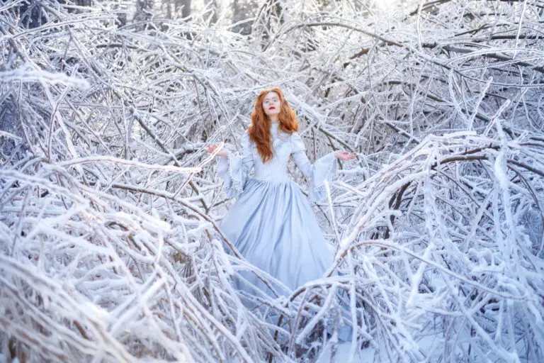 Red-haired woman in a pale blue gown surrounded by snow-covered branches in a winter forest.