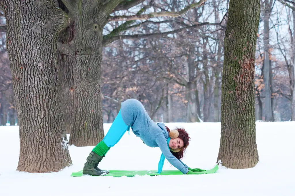 Woman practicing Downward-Facing Dog on a yoga mat in a snowy park, wearing colorful winter clothing.